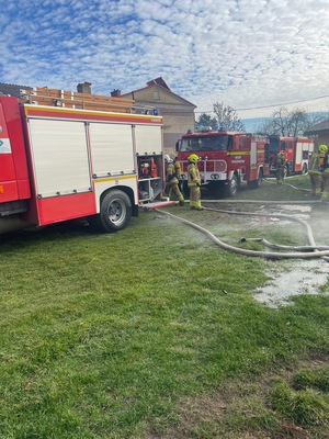 Trzy wozy strażackie przed płonącym budynkiem i strażacy.