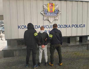 Odwróceni tyłem dwaj funkcjonariusze z zatrzymanym mężczyzną zakutym w kajdanki.