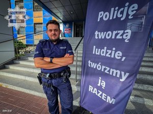 policjant stojący przy banerze z napisem &quot;Policję tworzą ludzie, tworzymy ja RAZEM&quot;, w tle schody