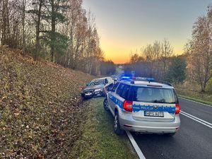 Na skraju drogi stoi radiowóz policyjny. Za nim w głębi samochód osobowy stojący częściowo w rowie.