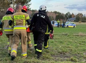 Strażacy i druhowie ochotnicy niosą nosze z odnalezioną kobietą, w tle radiowozy.
