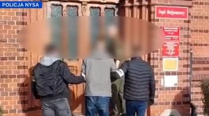 Dwaj policjanci prowadzą zatrzymanego mężczyznę w kierunku budynku sądu, zakutego w kajdanki.