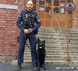 Policjant z psem służbowym stoi przy schodach wejściowych do budynku.