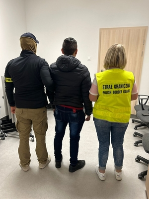 Funkcjonariuszka Straży Granicznej i policjant trzymają zatrzymanego mężczyznę.
