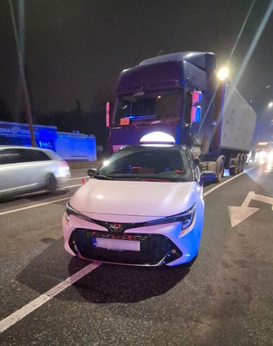 Na pierwszym planie samochód osobowy na drodze, za nim stoi samochód ciężarowy.