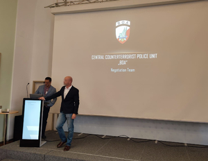 Dwaj mężczyźni stoją przy mównicy podczas wystąpienia w trakcie konferencji na tle ekranu, na którym wyświetlana jest prezentacja.