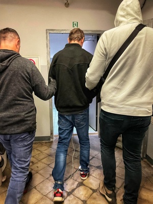 Dwaj policjanci prowadzą zatrzymanego mężczyznę zakutego w kajdanki zespolone.