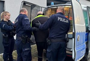 Policjantka i dwaj policjanci z zatrzymanym mężczyzną przy radiowozie.