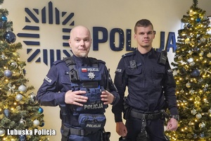 dwóch policjantów pozuje na tle napisu policja oraz przy dwóch choinach.