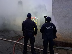 Stojący tyłem dwaj policjanci, przed nimi widać kłęby dymu i pożar.