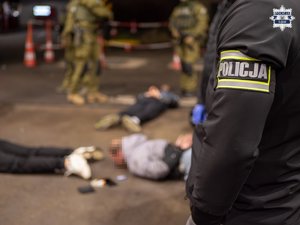 zdjęcie przedstawia rękę funkcjonariusza Policji, w drugim planie leżące na ziemi osoby.