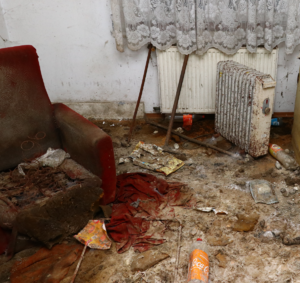 Wnętrze pustostanu. Po lewej stronie z niszczony fotel. N podłodze mnóstwo śmieci.