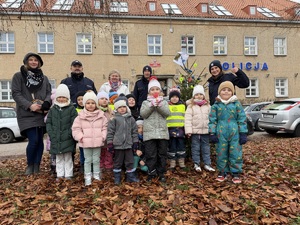 Policjanci oraz grupa małych dzieci z opiekunkami pozują do zdjęcia. W tle budynek komendy Policji.