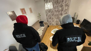 Dwaj stojący tyłem policjanci w kapturach na głowach i kurtkach z napisem Policja na plecach. Przed nimi widać porozkładane na stole i rozrzucone na kanapie banknoty o różnych nominałach.