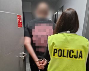 Policjantka w żółtej kamizelce z napisem Policja na plecach prowadzi zatrzymanego.