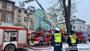 Pożar budynku, obok którego pracują służby m.in. Policji. Przy budynku wozy strażackie.
