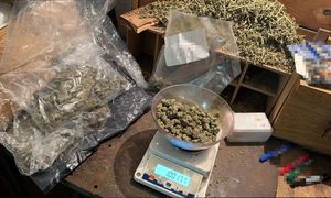 widać wagę, narkotyki w woreczku foliowym i zgrzewnicę. białe kryształki w worku foliowym, następnie dłoń w gumowej rękawiczce przekłada worki z marihuaną w kartonie. radiowóz, policjanta, który zakłada kajdanki zatrzymanemu, który leży na ziemi, następnie widok na pomieszczenie, w którym są paczki i worki z narkotykami, leżący luźno susz i półki na których są różne przedmioty. W pomieszczeniu panuje rozgardiasz.