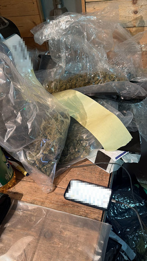 widać wagę, narkotyki w woreczku foliowym i zgrzewnicę. białe kryształki w worku foliowym, następnie dłoń w gumowej rękawiczce przekłada worki z marihuaną w kartonie. radiowóz, policjanta, który zakłada kajdanki zatrzymanemu, który leży na ziemi, następnie widok na pomieszczenie, w którym są paczki i worki z narkotykami, leżący luźno susz i półki na których są różne przedmioty. W pomieszczeniu panuje rozgardiasz.