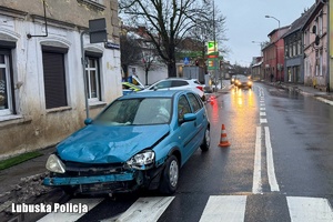 miasto, skrzyżowanie ze światłami, na pierwszym planie widać uszkodzony z przodu niebieski samochód, w tle widać biały samochód.