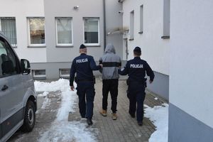 Dwaj policjanci prowadza zatrzymanego mężczyznę zakutego w kajdanki.