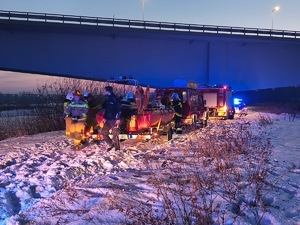 Wozy strażackie i łódź na brzegu rzeki obok mostu. Obok łodzi przyczepionej do jednego z pojazdów strażackich policjant i strażacy.