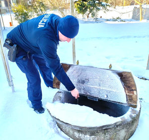 Dzielnicowy nabiera wodę ze studni trzymając łańcuch w ręku, teren dookoła pokryty jest  śniegiem.