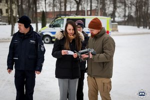 Policjant, obok niego stoi kobieta, która pokazuje kartkę operatorowi drona.