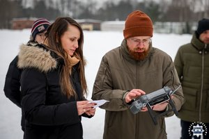 Policjant, obok niego stoi kobieta, która pokazuje kartkę operatorowi drona.