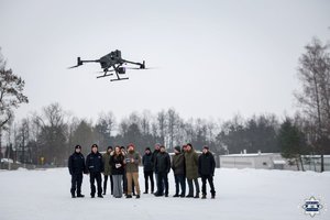 Nowoczesny dron ze skanerem 3D w powietrzu, na drugim planie widać grupę ludzi, w tym operatora drona i policjantów.