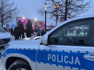 Na pierwszym planie bok radiowozu policyjnego,  w tle dwaj policjanci na koniach podczas patrolu i uczestnicy finału WOŚP.