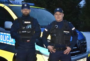 dwóch policjantów pozuje do zdjęcia w pełnym umundurowaniu. za nimi w tle stoi radiowóz.