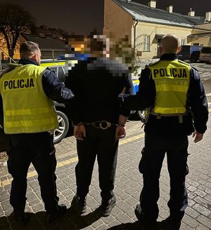 Dwaj policjanci w żółtych kamizelkach z napisem Policja na plecach prowadzą zatrzymanego.