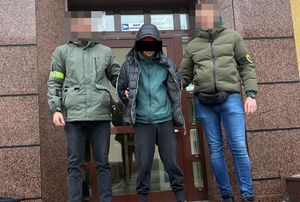 Dwaj policjanci z zatrzymanym mężczyzną stoją tyłem do wejścia do budynku.