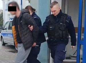 Policjant i policjantka prowadza zatrzymanego mężczyznę zakutego w kajdanki.