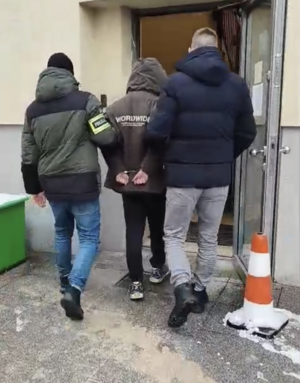 Dwaj funkcjonariusze prowadzą zatrzymanego mężczyznę zakutego w kajdanki  w kierunku budynku.