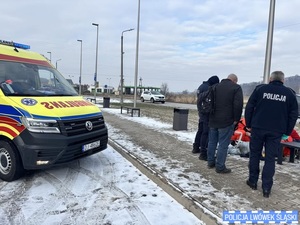 Dwaj policjanci i mężczyzna stoją obok ratowników medycznych udzielających pomocy mężczyźnie leżącemu na chodniku. Obok stoi karetka.
