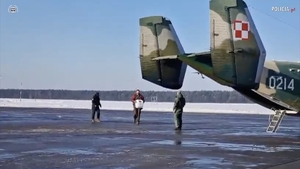 Ratownik medyczny niesie pojemnik z serem w kierunku wojskowego samolotu stojącego na płycie lotniska. Obok stoją lotnicy.