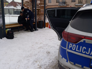 na pierwszym planie widać radiowóz z otwartymi drzwiami. w tle widać policjanta i mężczyznę siedzących na przystanku.