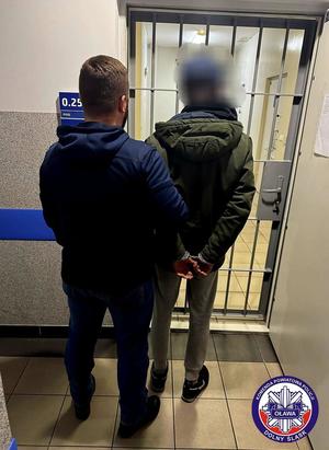 Policjant trzyma zatrzymanego mężczyznę zakutego w kajdanki. Stoją na korytarzu przed drzwiami z krat.