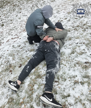 Policjant trzyma zatrzymanego mężczyznę leżącego na pokrytej śniegiem trawie.