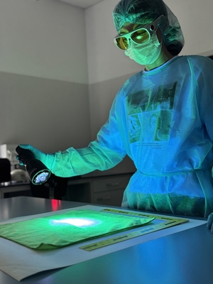 Osoba w fartuchu, w maseczce na twarzy i w okularach ogląda przedmiot leżący na blacie.