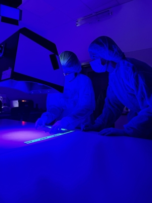 Dwie osoby ubrane w specjalne kombinezony, w maseczkach na twarzy i okularach oglądają przedmiot w świetle ultrafioletowym.
