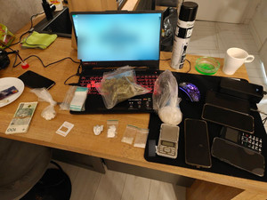 Na stole przed i obok laptopa leżą telefony komórkowe, środki odurzające w woreczkach, karta płatnicza i plik banknotów.