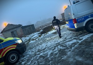 Policjant przy radiowozie, w głębi zabudowania.