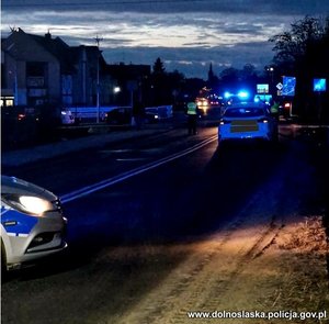 Miejsce zdarzenia. Zmrok. Ulica. Z lewej strony maska radiowozu policyjnego. W głębi na drodze stoi samochód osobowy, na środku drogi stoi policjant.