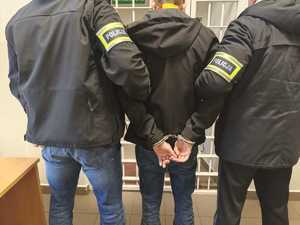 Dwaj funkcjonariusze prowadzą zatrzymanego mężczyznę zakutego w kajdanki.