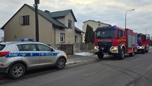 Przed budynkiem stoi zaparkowany policyjny radiowóz i wóz straży pożarnej.