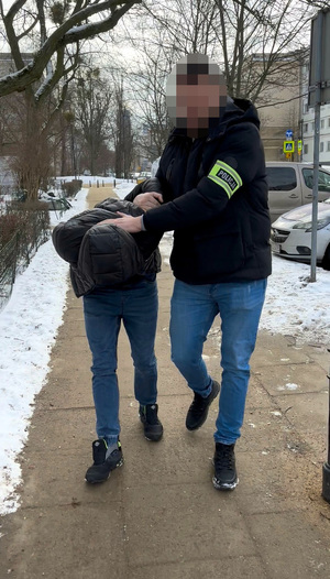 Funkcjonariusz prowadzi chodnikiem zatrzymanego mężczyznę zakutego w kajdanki.