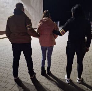 Funkcjonariusz i funkcjonariuszka trzymają zatrzymaną kobietę za ramiona.