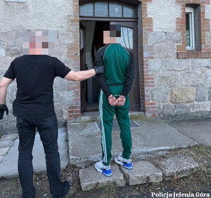 Funkcjonariusz przed wejściem do budynku trzyma zatrzymanego mężczyznę zakutego w kajdanki.